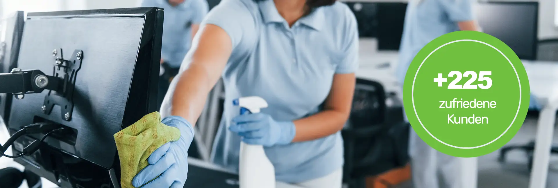 Büroreinigung - Kurzhaarige brünette Frau in einem hellblauen T-Shirt, die einen Monitor auf einem Schreibtisch mit einem Lappen säubert - Clean Team Reinigung von Büros