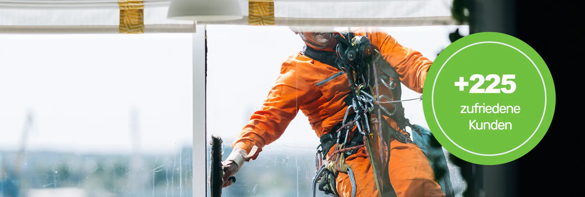 Gebäudereinigung Köln - Mann in orangenfarbener Arbeitskleidung ist durch Seile gesichert und putzt in großer Höhe eine Fensterfront von außern - Clean Team