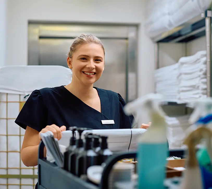 Hotelreinigung - Lächelnde Frau mit blondbraunen Haaren steht in einem Wäscheraum eines Hotel, die in einem Stahl-Regal weiße handtüscher beinhalten. Sie schiebt einen Wagen mit Reinigungsutensilien - Clean Team Reinigung von Hotels